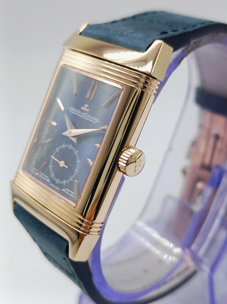 K&ouml;nnte beinhalten: Eine rechteckige, goldfarbene Armbanduhr mit dunkelblauem Zifferblatt und kleiner Sekunde. Die Uhr hat goldene Zeiger und Markierungen, eine Krone mit Logo und ein dunkelblaues Lederarmband. Die Uhr wird auf einem transparenten St&auml;nder pr&auml;sentiert.