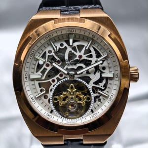 Puede incluir: Un reloj de pulsera de oro rosa con esfera esqueletizada que revela complejos componentes mecánicos. El reloj cuenta con una correa de piel negra, manecillas luminiscentes y una jaula de tourbillon dorada en la posición de las seis. La esfera muestra el texto Vacheron Constantin Geneve y Swiss Made.