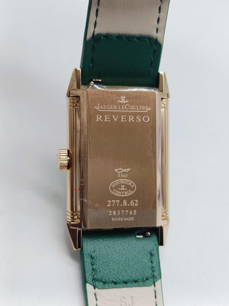 Puede incluir: Reloj Jaeger-LeCoultre Reverso dorado con correa de cuero verde. La esfera del reloj muestra el nombre de la marca y los detalles del modelo. La parte trasera del reloj tiene el texto "3 bar", "1000 hours control" y "Swiss Made".