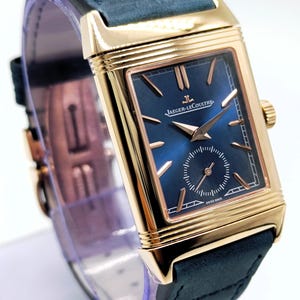 K&ouml;nnte beinhalten: Eine rechteckige Armbanduhr in Goldoptik mit einem marineblauen Zifferblatt und einem marineblauen Lederarmband. Das Zifferblatt der Uhr hat goldfarbene Zeiger und Markierungen, mit dem Markennamen "JAEGER-LECOULTRE". Eine Luxusuhr.