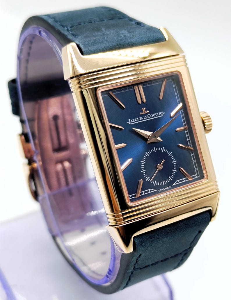 K&ouml;nnte beinhalten: Eine rechteckige Armbanduhr in Goldoptik mit einem marineblauen Zifferblatt und einem marineblauen Lederarmband. Das Zifferblatt der Uhr hat goldfarbene Zeiger und Markierungen, mit dem Markennamen "JAEGER-LECOULTRE". Eine Luxusuhr.