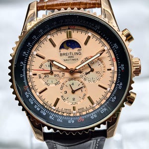 May include: A Breitling 1884 Navitimer watch with a rose gold-tone case and a light copper-coloured dial. The watch features a moon phase indicator, multiple sub-dials for day, date, and month, a black outer slide rule bezel, and a brown leather strap.