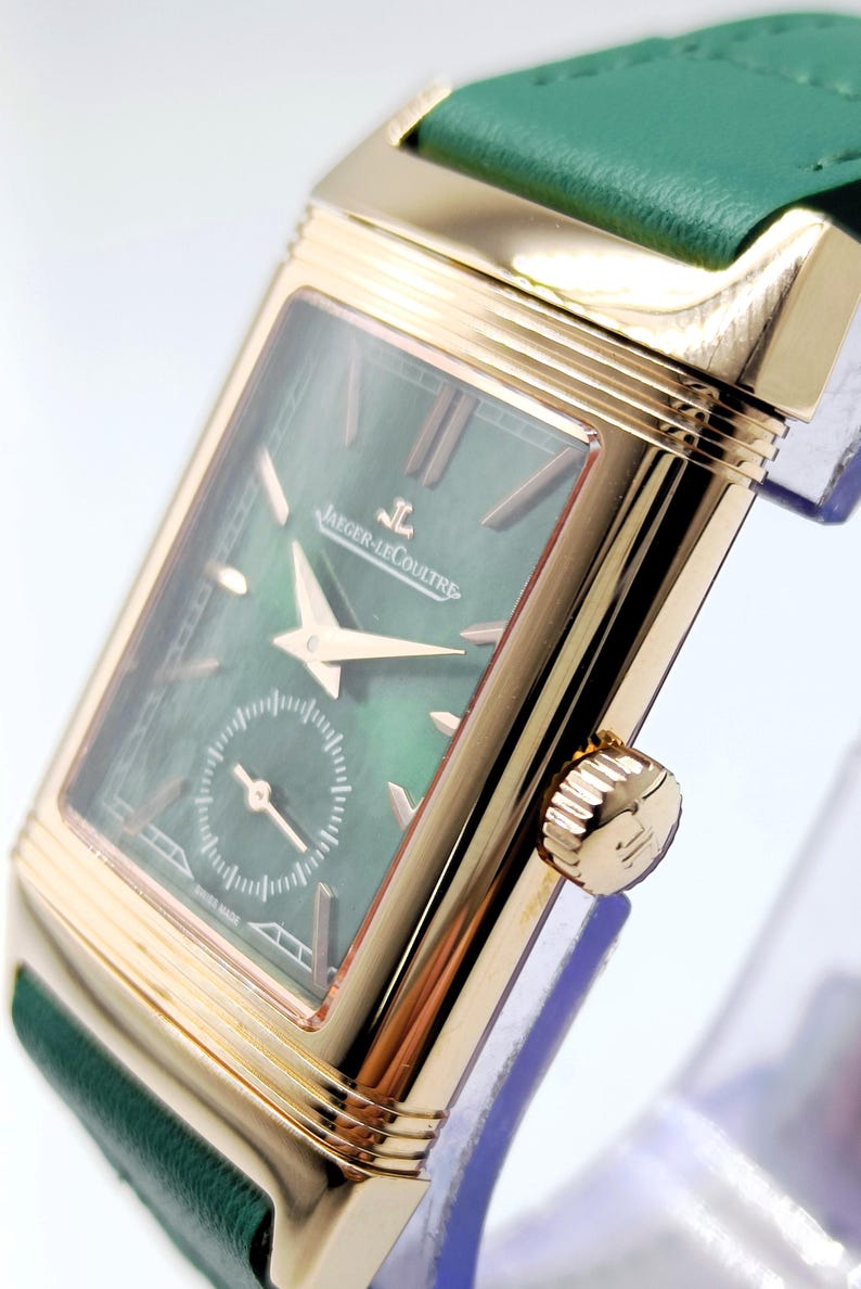Puede incluir: Un reloj de pulsera rectangular dorado con esfera verde y correa de cuero verde. La esfera del reloj presenta la marca "Jaeger-LeCoultre" y un peque&ntilde;o dial de segundos. El reloj tiene agujas e indicadores de hora dorados.