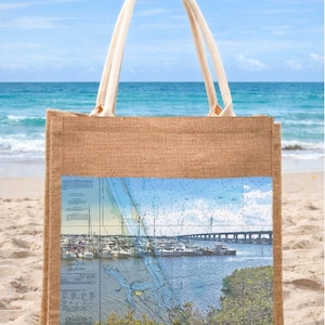 May include: A jute tote bag with cream-colored handles, resting on a sandy beach. The bag features a rectangular print of a nautical chart with a harbor scene, a bridge, and the text "Stuart, FL" with coordinates.