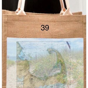 Puede incluir: Un bolso tote de yute de tono natural con asas color crema. El bolso presenta un parche rectangular con un mapa de Cape Cod, Massachusetts, y el texto "Cape Cod" con coordenadas. El número "39" está impreso encima del parche.