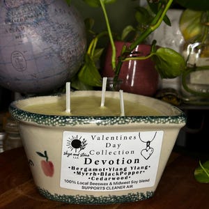 Puede incluir: Una vela cuadrada de color crema con tres mechas, etiquetada como "Devotion" de la "Colección de San Valentín". La vela tiene un borde verde e ilustraciones de manzanas rojas. La etiqueta enumera los ingredientes y apoya el aire más limpio.