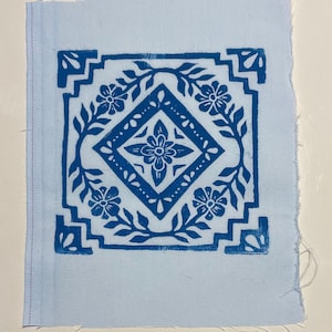 Puede incluir: Un cuadrado de tela azul claro con un estampado en bloque azul. El estampado presenta una forma de diamante central con un diseño floral, rodeado por un borde floral. La tela tiene un fondo azul claro.