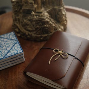 May include: A brown leather journal secured with a thin black elastic band featuring a gold-toned metal bow charm. The journal has a smooth, matte finish and contains white paper pages visible at the edge. The cover is plain with no visible text.