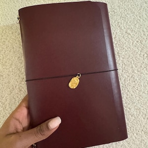 May include: A dark burgundy leather notebook cover featuring a thin black elastic band wrapped horizontally around the center. A small, oval-shaped gold-toned metal charm with an embossed floral design is attached to the elastic band.