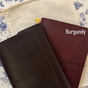 May include: Two leather journals, one chocolate brown and one burgundy, are displayed on a floral patterned surface. The burgundy journal has the word "Burgundy" printed on it. A cream-coloured drawstring bag is in the background.