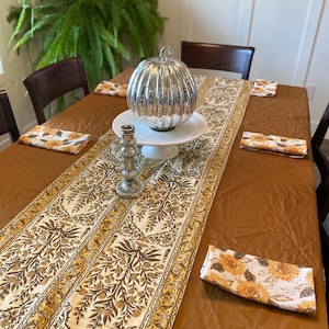 May include: A dining table set for a fall gathering. The table is covered with a brown tablecloth and a floral runner. A silver pumpkin sits on a white cake stand, with matching sunflower napkins.