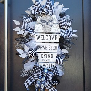 Könnte beinhalten: Ein Halloween-Kranz mit einem Totenkopf-Design und dem Text "WELCOME, WE'VE BEEN DYING TO MEET YOU." Der Kranz hat schwarz-weiße Bänder, dekorative Kugeln und eine Schleife. Der Kranz ist mit weißen Blättern und schwarzen, farnartigen Akzenten verziert.
