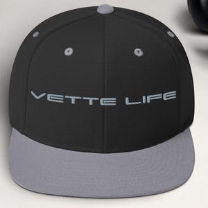 May include: A black and gray baseball cap with the text "VETTE LIFE" embroidered in gray. The cap has a flat brim and a structured fit. The hat is a classic style, suitable for everyday wear.
