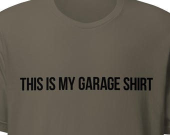 May include: Olive green t-shirt with the text "THIS IS MY GARAGE SHIRT" printed in black across the chest. The shirt has short sleeves and a crew neck.