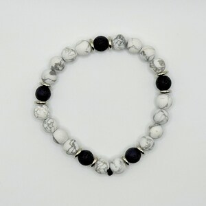 May include: A beaded bracelet featuring white and gray marbled beads interspersed with black lava beads. The bracelet is strung on a black cord and accented with silver-colored metal spacers. The bracelet is designed for wrist wear.
