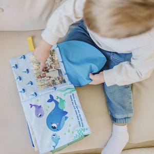 Puede incluir: Un libro para bebés azul con un diseño de ballenas y veleros. El libro tiene un bolsillo con una foto en su interior. El texto del libro dice: "Seres queridos cerca y lejos. Su amor por mí nunca vacilará."