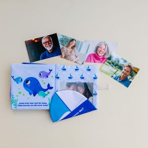 May include: A soft baby photo album with a blue and white whale design and the text "Loved ones near and far away. Their love for me will never sway!" Several photos are displayed around the album.