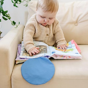 May include: A baby is seated on a beige sofa, looking at a colorful soft book. The book has various illustrations and text. A blue, round, soft object is in the foreground. The baby is wearing a beige cardigan.