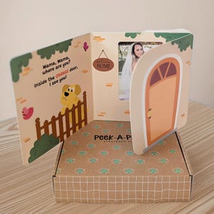 May include: A Peek-A-P Storybook album with an orange door design. The open book displays a photo and text: "Mama, Mama, where are you? Inside the ORANGE door, I see you!" A brown box is beneath the book.