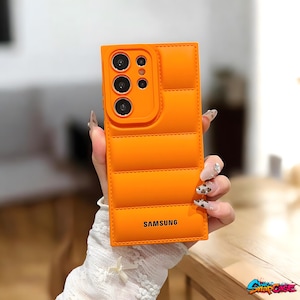 May include: Vibrant orange phone case with a quilted pattern, camera aperture, and the "SAMSUNG" brand. The rectangular case is held by a person. The case is designed for a mobile phone.