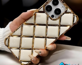 May include: A white and gold phone case with a quilted design. The case has a gold frame and a white, square-patterned back. The phone's camera lenses are visible. The case is held by a person.