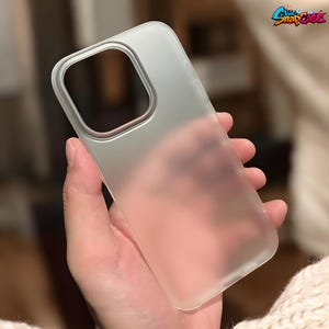 Puede incluir: Una funda de teléfono transparente esmerilada sostenida en una mano. La funda tiene un recorte para la cámara y un logotipo en la esquina superior derecha. La funda está hecha de un material translúcido.