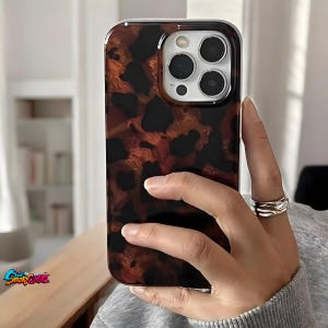 Op de afbeelding: Een telefoonhoesje met schildpadpatroon en een doorzichtige rand. De hoes heeft een bruin en zwart gevlekt ontwerp. De telefoonhoes wordt in een hand gehouden en de cameralenzen van de telefoon zijn zichtbaar. Het logo "Chic SnapCase" staat linksonder.