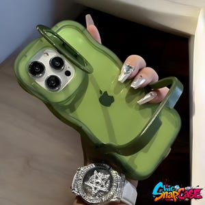 Könnte beinhalten: Eine transparente grüne Handyhülle mit Griff und erhöhtem Ring. Die Hülle hat einen Ausschnitt für die Kamera des Telefons und das Apple-Logo. Die Hülle wird von einer Person gehalten, die eine Uhr mit weißem Armband und einem diamantbesetzten Zifferblatt trägt.