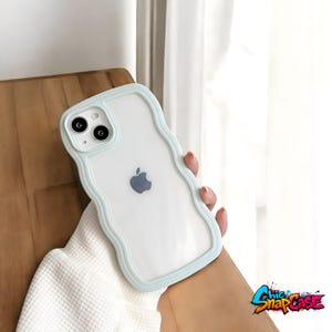 May include: A clear iPhone case with a wavy, light blue border. The case has a transparent back, showcasing the phone's design, including the Apple logo and camera lenses. The case is held by a person wearing a white sweater. The text "Chic SnapCase" is visible.
