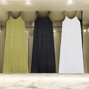 May include: Three long, sleeveless dresses with thin straps. The dresses are in olive green, black, and white. The dresses have a textured, vertical ribbed pattern. The dresses are hanging in a well-lit space.