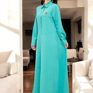 Peut inclure: Longue robe turquoise avec col chemise et manches longues. La robe présente une fermeture boutonnée sur le devant et une petite broche décorative près du col. La robe a une jupe longue et fluide.