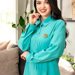 Peut inclure: Une robe chemise turquoise à manches longues et col chemise. Une broche dorée est épinglée sur la poitrine. La robe est assortie d'un collier et de boucles d'oreilles dorés. La robe est faite d'un tissu lisse et fluide.