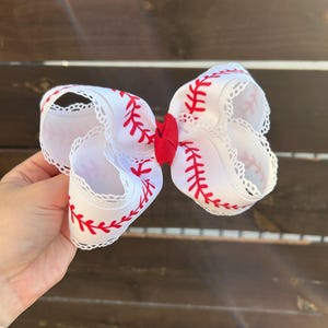 May include: A white hair bow with red baseball stitching and a red center. The bow is made of a wide, decorative ribbon with a scalloped edge. The baseball-themed bow is held in a hand.