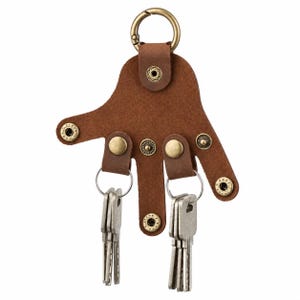 Peut inclure: Un organisateur de clés en cuir marron en forme de main à cinq doigts, doté de boutons-pression et de rivets en métal couleur laiton. Un mousqueton circulaire en métal est fixé au sommet, et deux anneaux en métal argenté retiennent des clés.