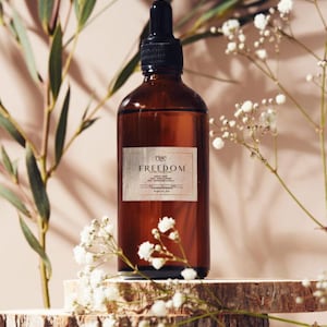 Op de afbeelding: Een bruine glazen fles met een zwarte druppelkap, met het label "rise FREEDOM". De fles staat op een houten plank, omringd door witte bloemen en groene bladeren. De achtergrond is een zachte, neutrale kleur.