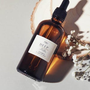 Può includere: Una bottiglia di vetro marrone con un contagocce nero, etichettata "rise DEEP". La bottiglia è angolata su una superficie di colore chiaro, con un pezzo di legno e piccoli fiori bianchi sullo sfondo. La bottiglia contiene un liquido.