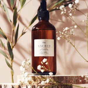 May include: A brown glass bottle with a black dropper, labeled "Sacred" with white text. The bottle sits on a wooden platform, surrounded by white flowers and green foliage. The product is a diffuser.