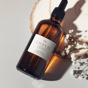 Pode incluir: Uma garrafa de vidro castanho com um conta-gotas preto, rotulada "Eternal" com texto branco. A garrafa está angulada sobre uma fatia de madeira com flores brancas. O rótulo também inclui a marca "rise".