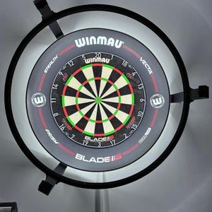 Op de afbeelding: Een Winmau Blade 6 dartbord met een zwarte rand en ingebouwde verlichting. Het dartbord heeft een zwart-grijze voorkant met rode, groene en witte details. Het Winmau-logo is zichtbaar.