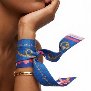 Könnte beinhalten: Ein blau und dunkelblau gestreiftes Seidenband, das um ein Handgelenk gebunden ist, mit einem goldenen Seilmuster, rosa Blumenakzenten, einem kleinen gelben Schmetterling und dem deutschen Text Sei stets behend und wende dich dem Guten zu.