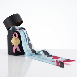 Könnte beinhalten: Eine schwarze zylindrische Geschenkbox mit abgenommenem Deckel, verziert mit einem rosa Band und einem goldenen Wachssiegel. Ein hellblaues Tuch mit weißem Karomuster, schwarzen Ovalen und roten Herzakzenten liegt davor, mit dem Text Glaube an die Liebe.