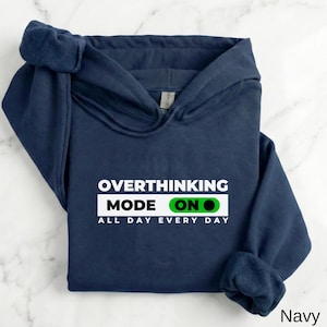 May include: A navy blue hooded sweatshirt featuring white text that reads OVERTHINKING MODE ON ALL DAY EVERY DAY. The word ON is inside a green toggle switch graphic. The hoodie has long sleeves, a front pocket, and a drawstring hood.