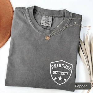 May include: A charcoal grey short-sleeved t-shirt with a crew neckline. The shirt features a white graphic on the lower right side depicting a shield with the text "PRINCESS SECURITY" and three stars. A gold-toned pendant necklace rests on the collar.