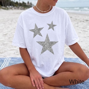 以下が含まれることがあります： 前面に3つの星がプリントされた白い半袖Tシャツ。星は質感のあるきらめくシルバーのグリッター仕上げで、中央に大きな星が1つ、その上にサイズの異なる小さな星が2つ配置されています。