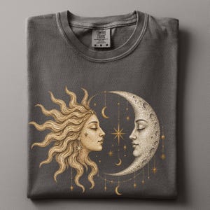 以下が含まれることがあります： チャコールグレーのTシャツで、太陽と三日月が向かい合っているゴールドトーンのグラフィックが描かれています。太陽には流れるような光線と人の顔があり、月にはクレーターのある表面と人の顔があります。小さな星や月が下にぶら下がっています。