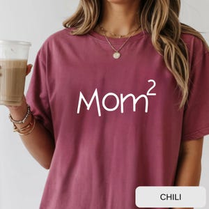 Könnte beinhalten: Ein weinrotes T-Shirt mit dem Wort "Mom²" in Weiß, ein verspieltes Design. Das Shirt hat einen Rundhalsausschnitt und kurze Ärmel. Das Wort "CHILI" ist auf einem weißen Rechteck am unteren Bildrand aufgedruckt.