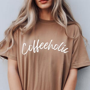 May include: A tan t-shirt featuring the word "Coffeeholic" in white cursive script. The top has short sleeves and a round neck. The model has long blonde hair.