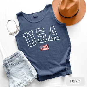 May include: A navy blue sleeveless tank top featuring the white block-letter text USA and a small American flag graphic. The garment is displayed alongside a tan fedora hat, a silver-toned floral necklace, and light-wash denim shorts.