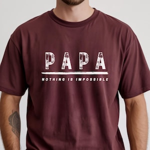 May include: A maroon t-shirt with the word "PAPA" in distressed white letters above the phrase "NOTHING IS IMPOSSIBLE". The shirt has a crew neck and short sleeves.