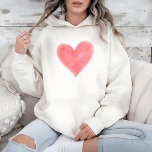 May include: Cream-colored hoodie featuring a large, watercolor-style pink heart on the front. The hoodie has a drawstring hood and a kangaroo pocket. The person is wearing light blue jeans.